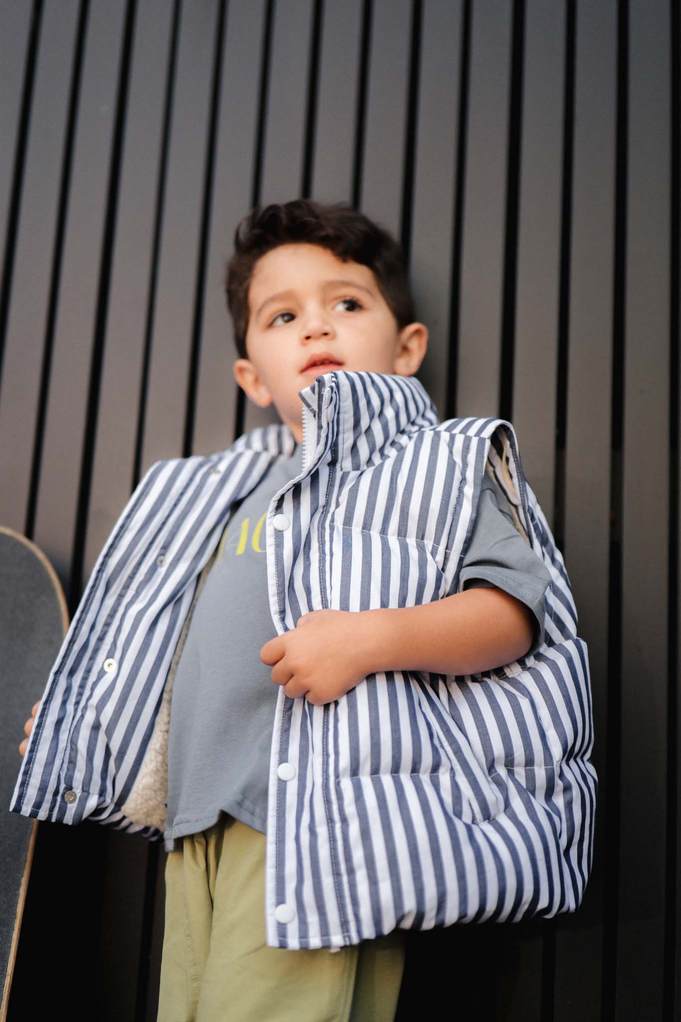 Child wearing a striped puffer vest and holding a skateboard against a dark background