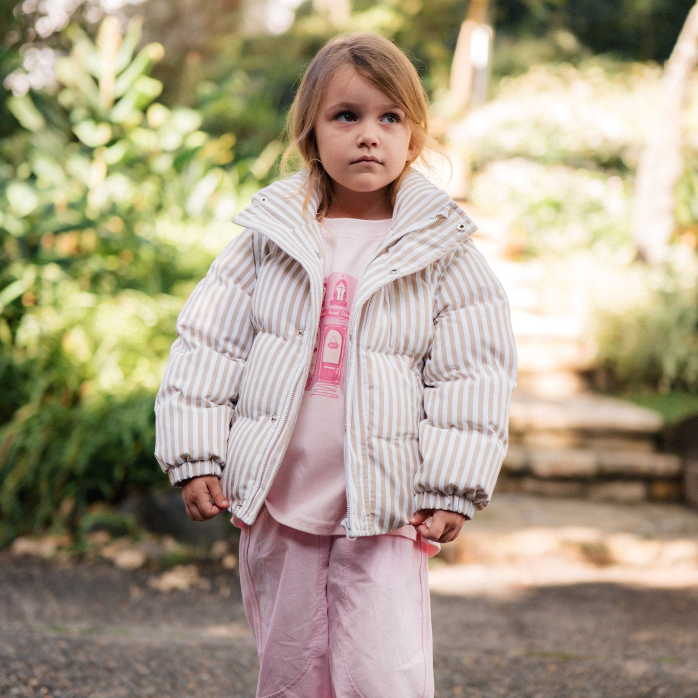 Child wearing a white puffer jacket and pink outfit standing outdoors with greenery in the background