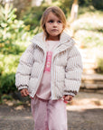 Child wearing a white puffer jacket and pink outfit standing outdoors with greenery in the background