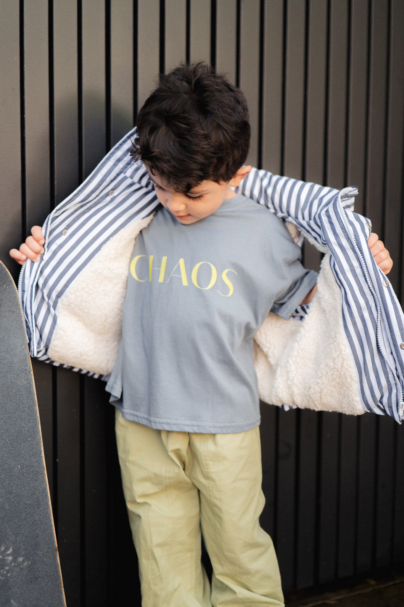 Child wearing a shirt with 'CHAOS' text, holding a striped jacket over their shoulders against a dark background.
