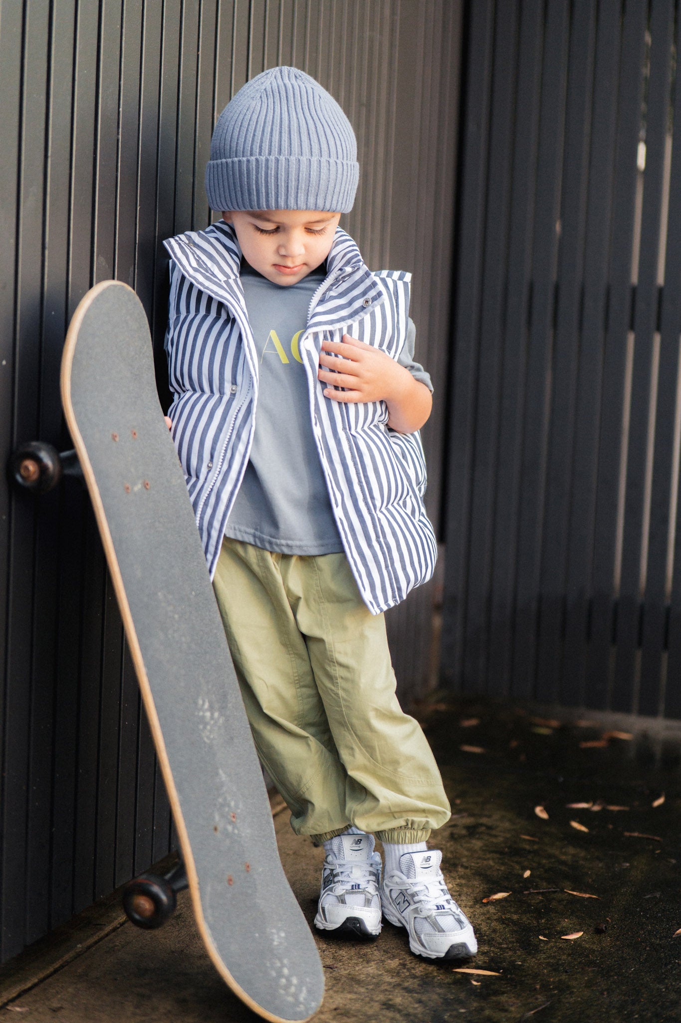 Child holding a skateboard, wearing a blue beanie, striped vest, and green pants against a dark background.