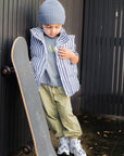 Child holding a skateboard, wearing a blue beanie, striped vest, and green pants against a dark background.