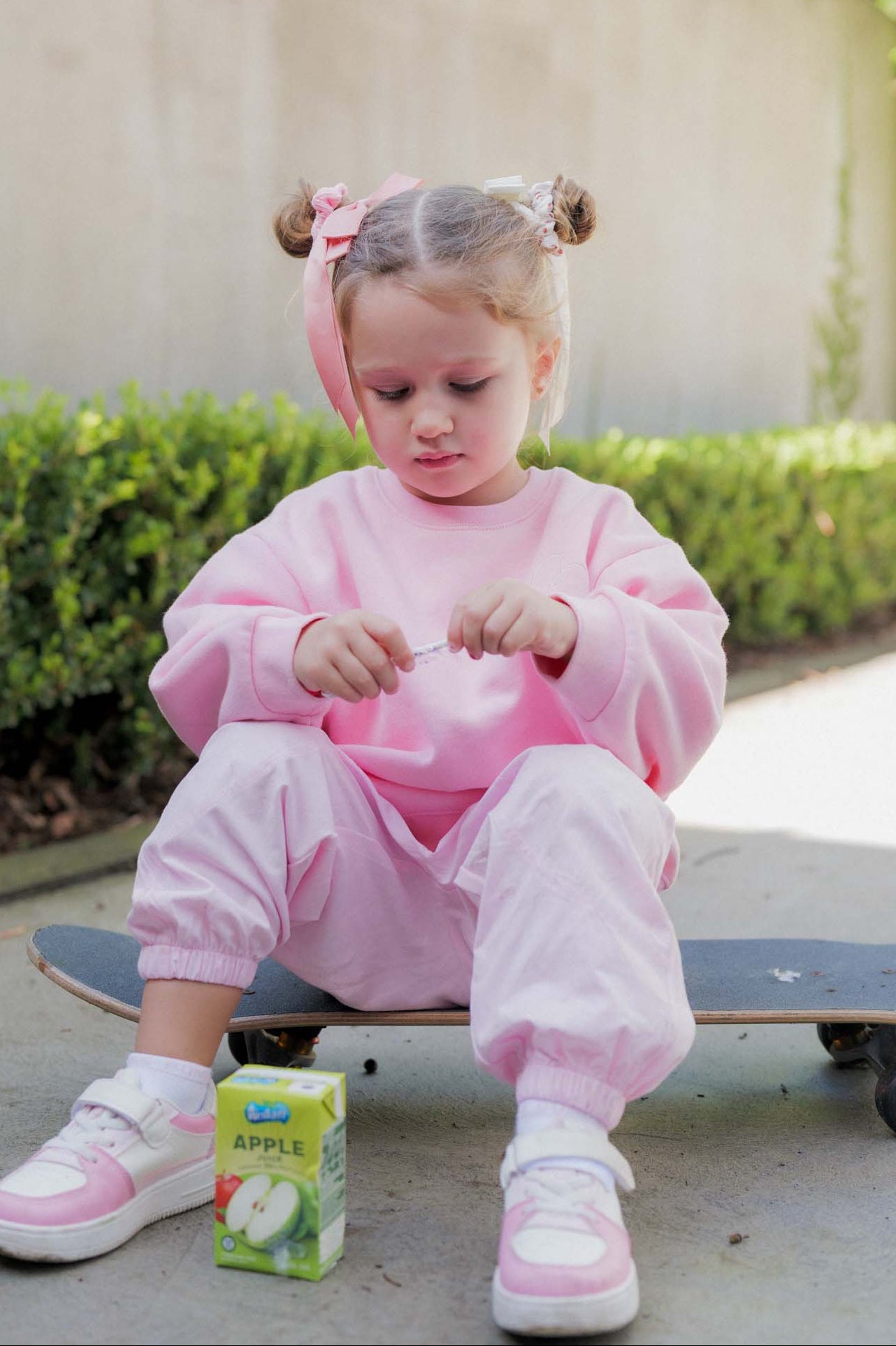 Child in pink outfit sitting on a skateboard with a juice box