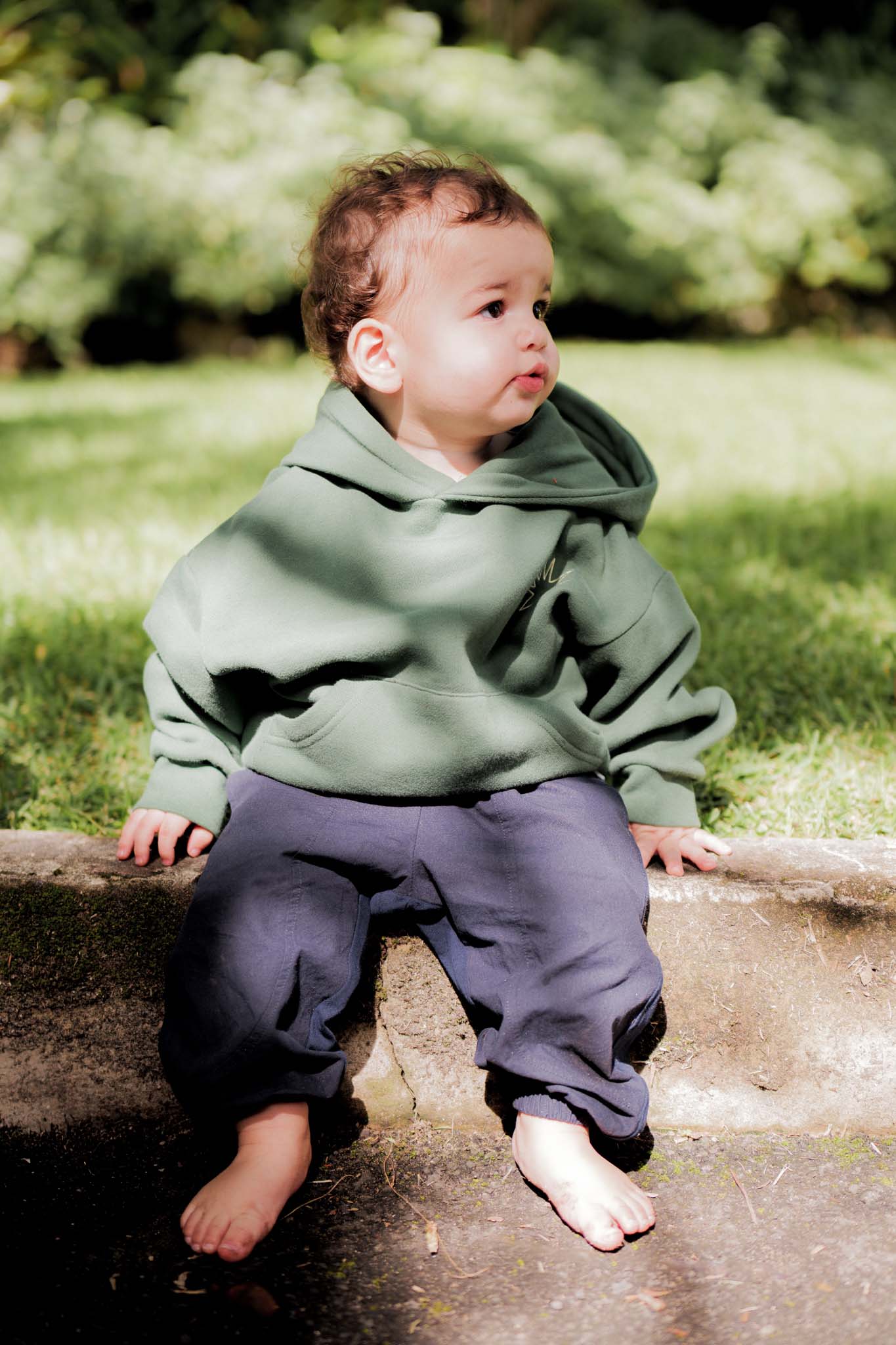 Child wearing a green hoodie and dark pants sitting on a log outdoors.