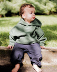 Child wearing a green hoodie and dark pants sitting on a log outdoors.