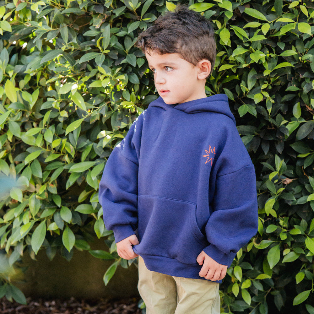 Child wearing a blue hoodie and olive pants standing in front of green foliage.
