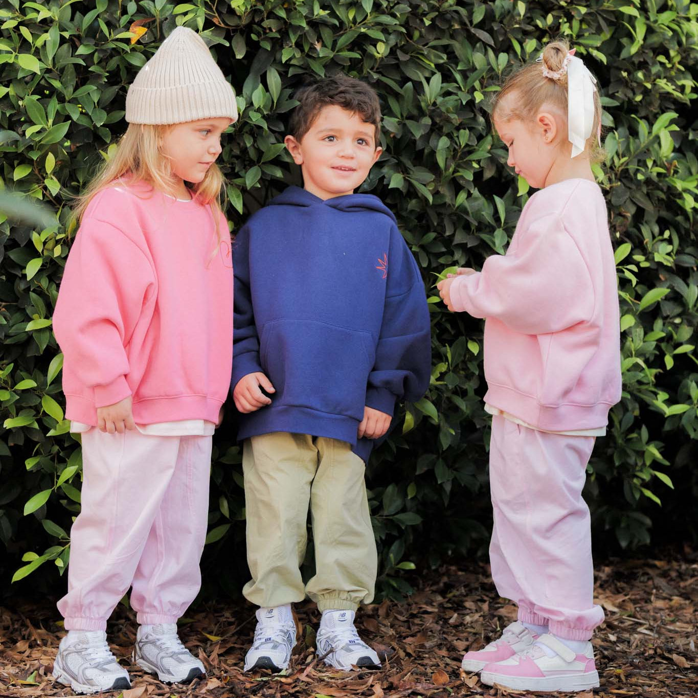 Three children in matching pink and blue outfits standing in front of a green bush.