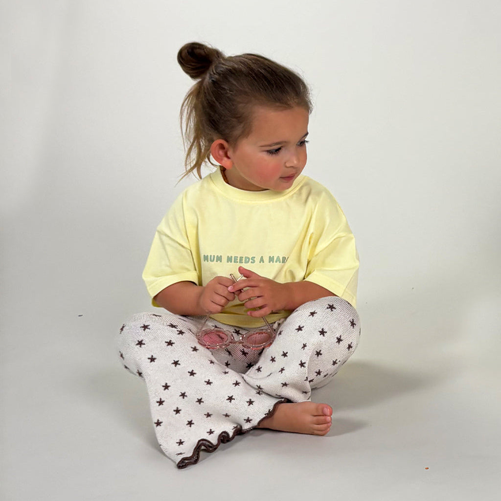 A toddler girl sitting cross-legged on a light gray studio floor, wearing a pale yellow graphic T-shirt with the text 'RUN NEEDS A MARGI' and white pants with a black star pattern.