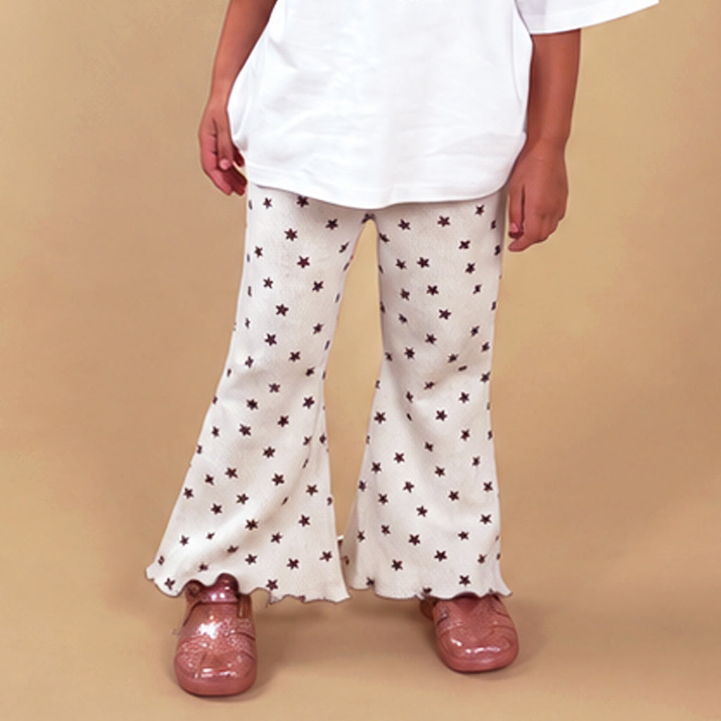 A child wearing cream flared pants with a dark brown star pattern, a white t-shirt, and pink shoes, standing against a solid beige background.