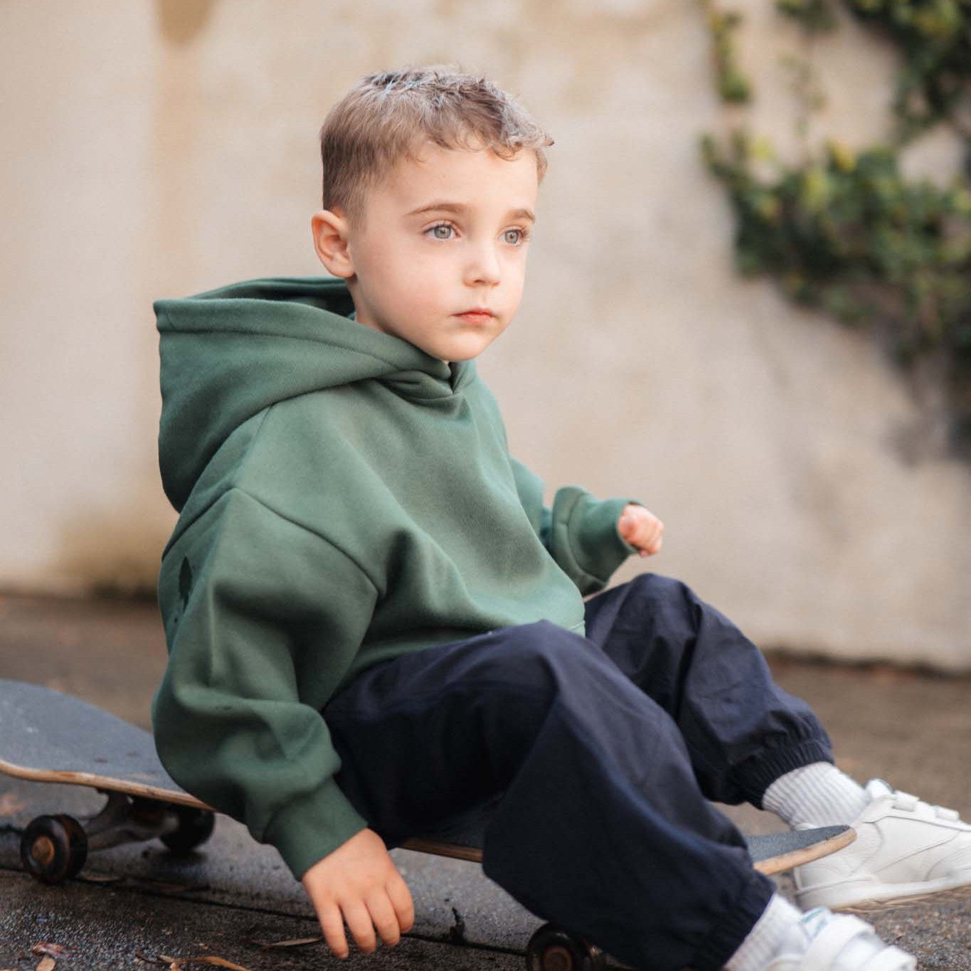 Child sitting on a skateboard wearing a green hoodie and dark pants.