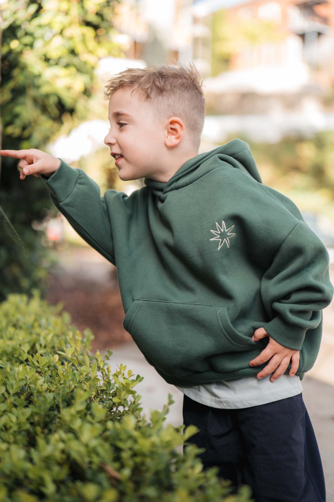 Child wearing a green hoodie with a star design, standing outdoors near greenery.