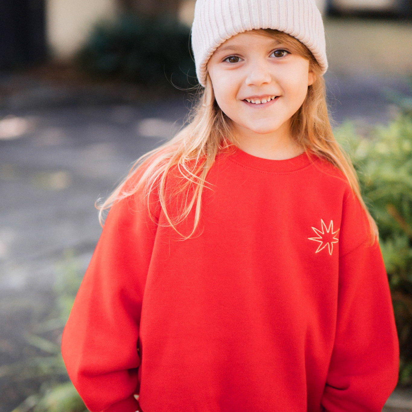 Young girl wearing a red sweatshirt with a logo and a white beanie outdoors.
