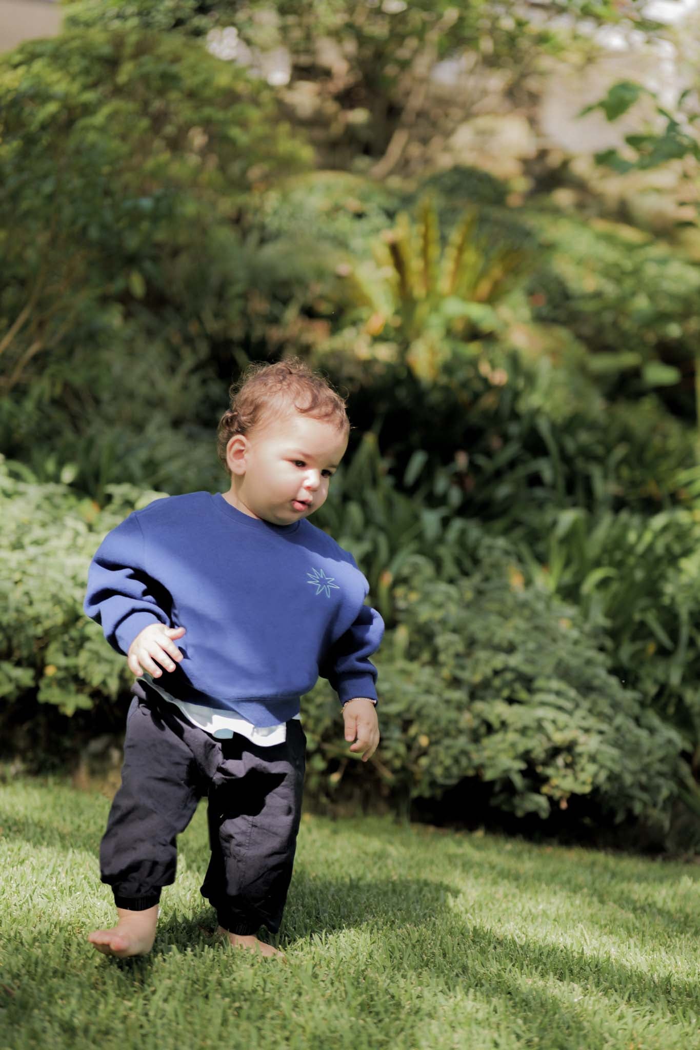 Child in a blue sweater and dark pants standing on grass with greenery in the background