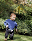 Child in a blue sweater and dark pants standing on grass with greenery in the background