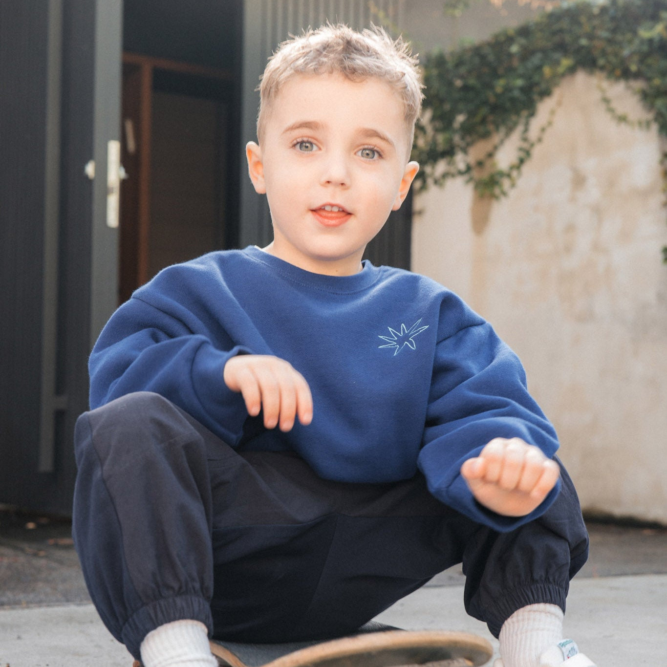 Child sitting on a skateboard wearing a blue sweatshirt and dark pants.