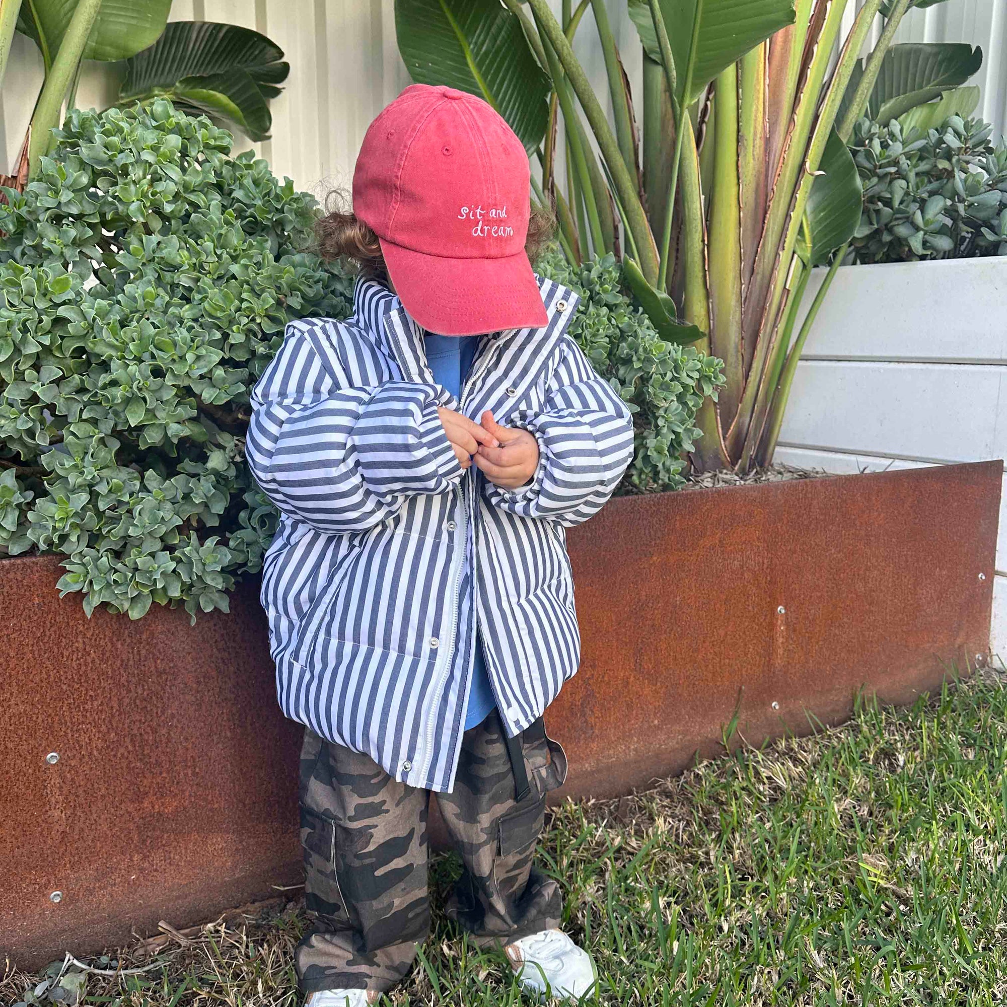 Child wearing a red cap and striped shirt standing in front of plants