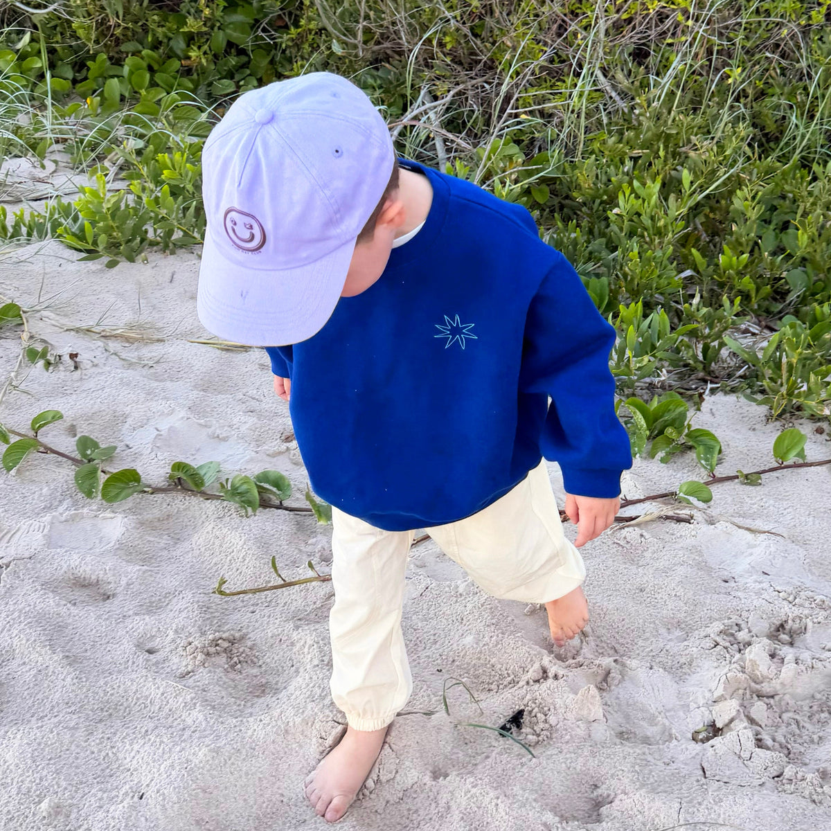A toddler walking barefoot on sand near green bushes, wearing a royal blue sweatshirt, cream pants, and a light lavender baseball cap.