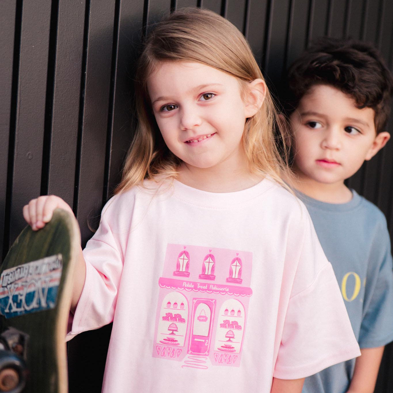 Two children, a girl holding a skateboard and a boy in the background, against a dark background.