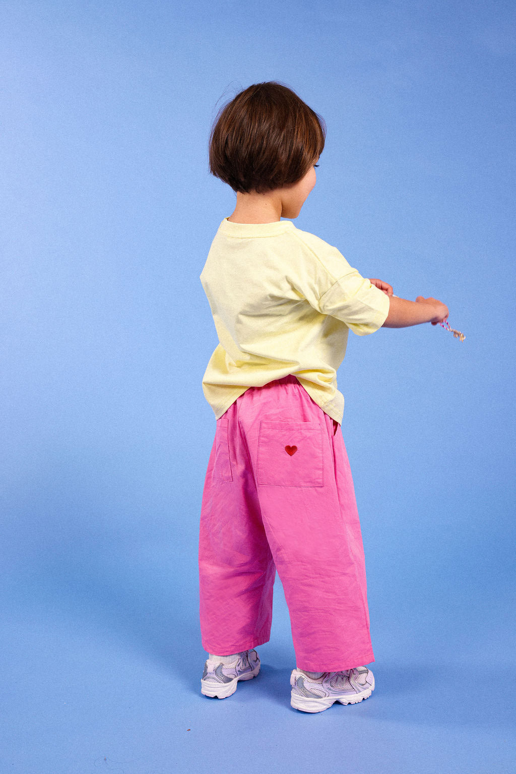 A toddler standing with their back to the camera, wearing a pale yellow t-shirt, bright pink wide-leg pants with a small red heart on the pocket, and white sneakers, set against a solid blue studio background.