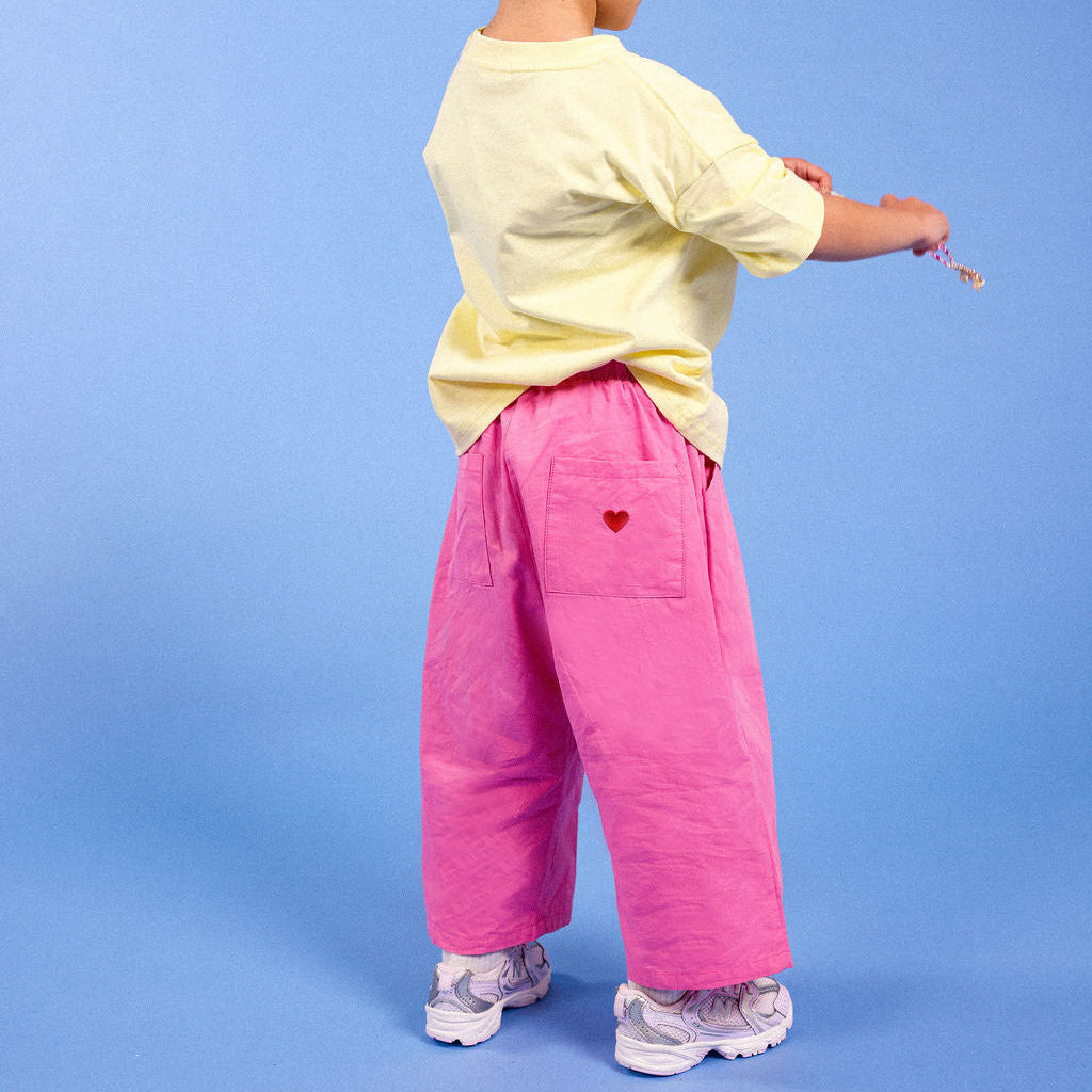 A toddler standing with their back to the camera, wearing a pale yellow t-shirt, bright pink wide-leg pants with a small red heart on the pocket, and white sneakers, set against a solid blue studio background.