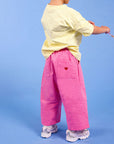 A toddler standing with their back to the camera, wearing a pale yellow t-shirt, bright pink wide-leg pants with a small red heart on the pocket, and white sneakers, set against a solid blue studio background.