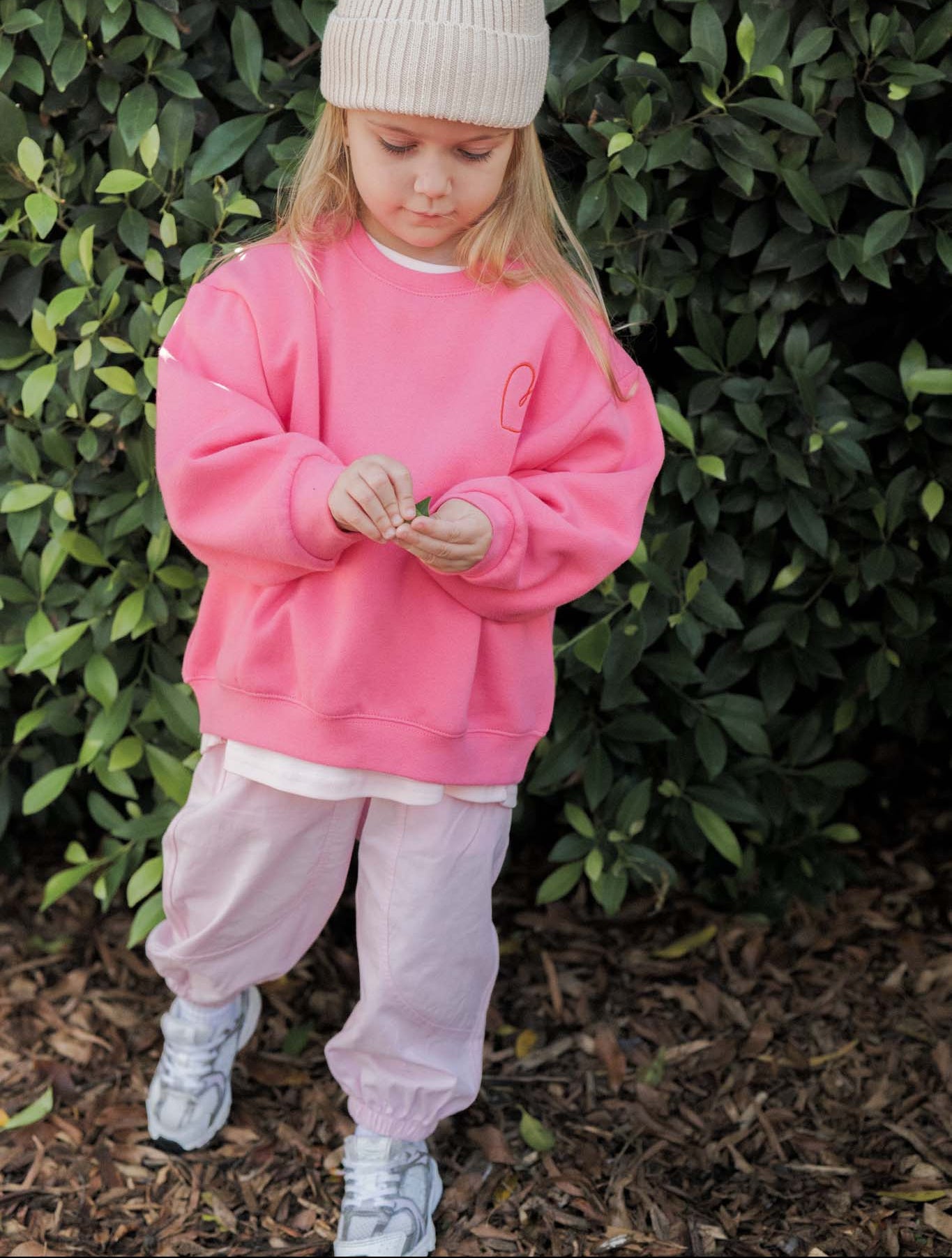 Child wearing a pink sweatshirt and light-colored pants standing in front of green foliage.