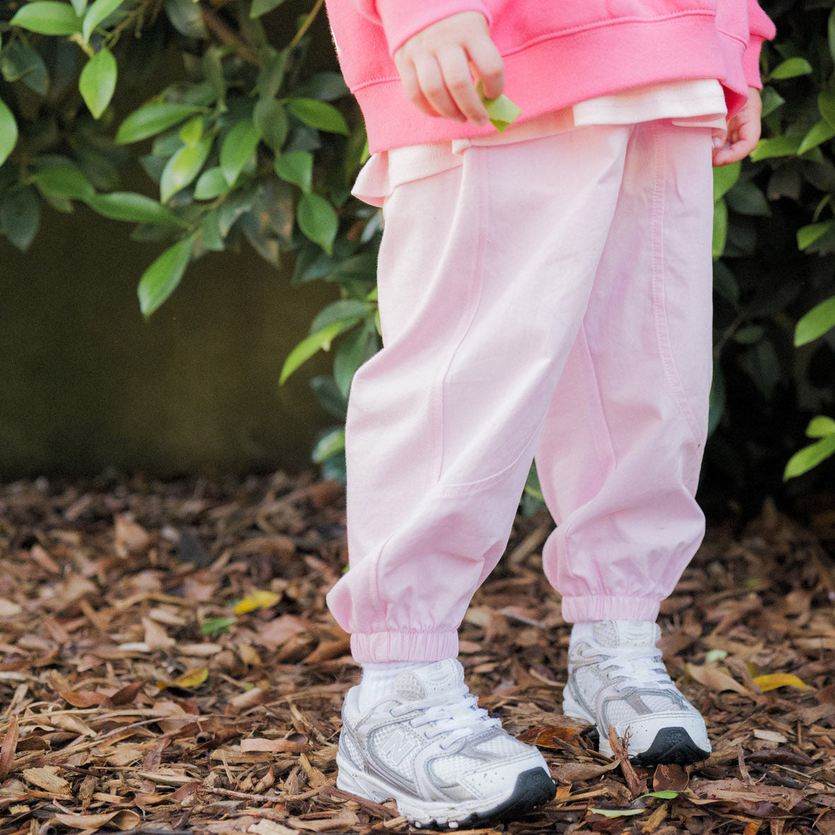 Person wearing a pink outfit with white shoes standing on mulch with greenery in the background