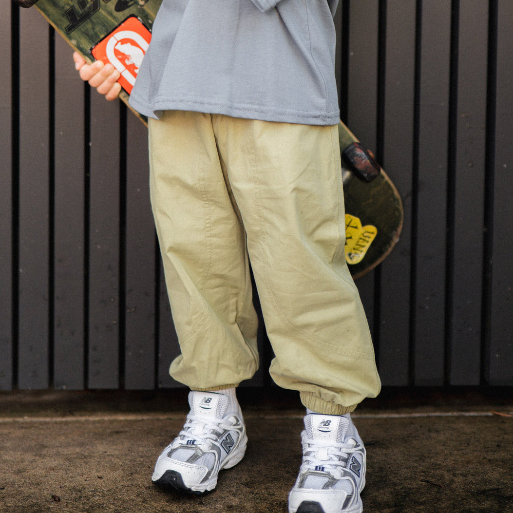 Person wearing olive pants and white sneakers holding a skateboard against a dark background