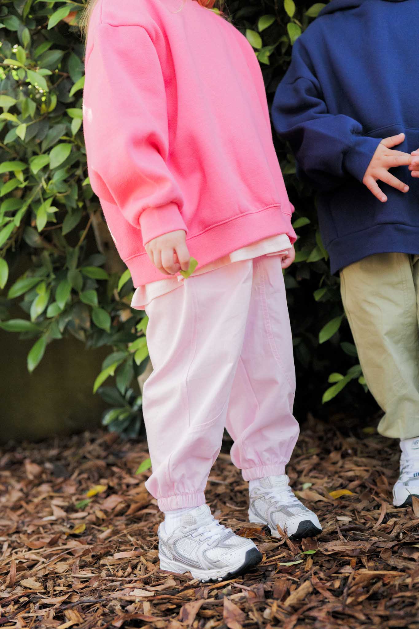 Two children wearing colorful outfits standing on mulch with greenery in the background