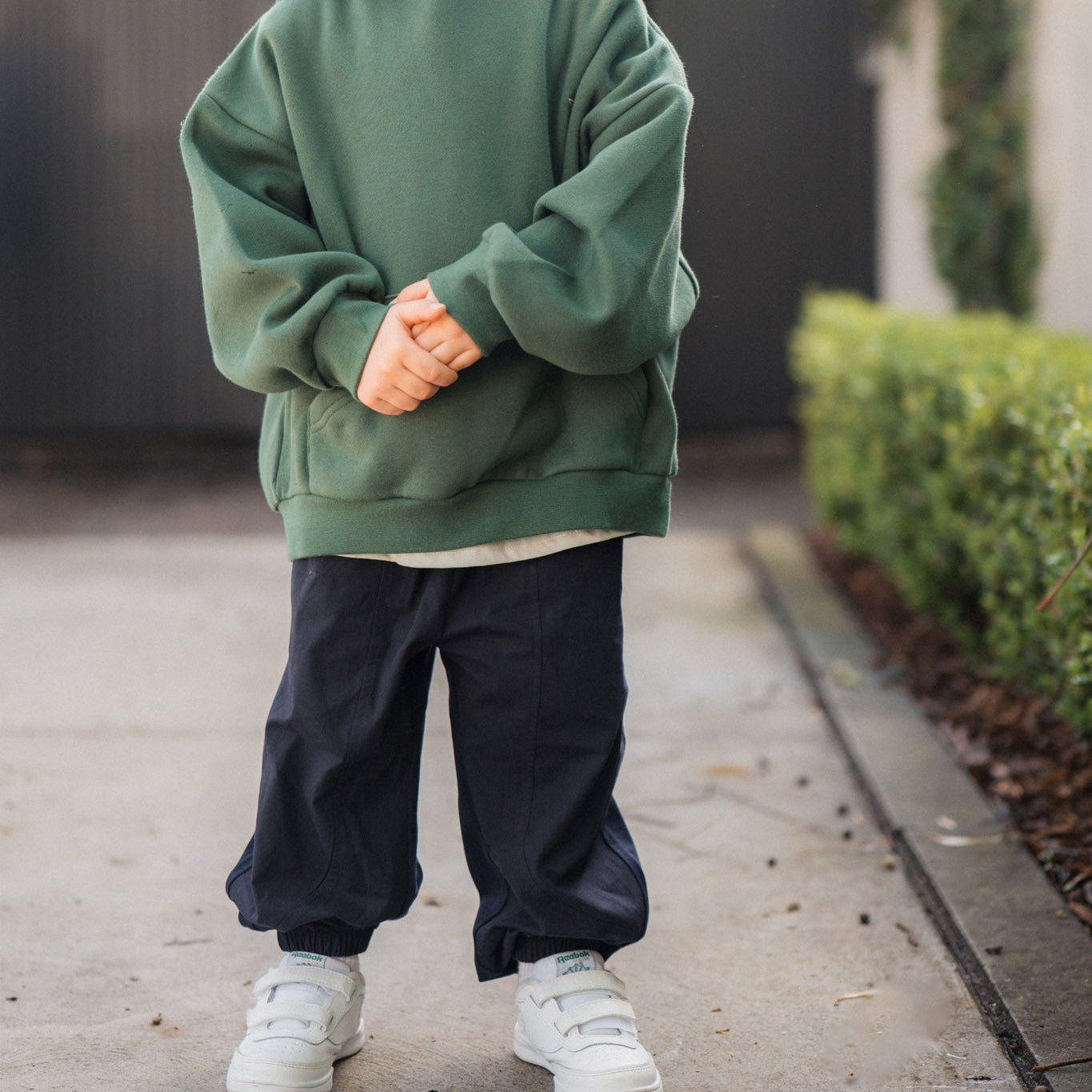 Child wearing a green hoodie and dark pants standing on a sidewalk.