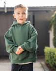 Child wearing a green hoodie and dark pants standing on a sidewalk.