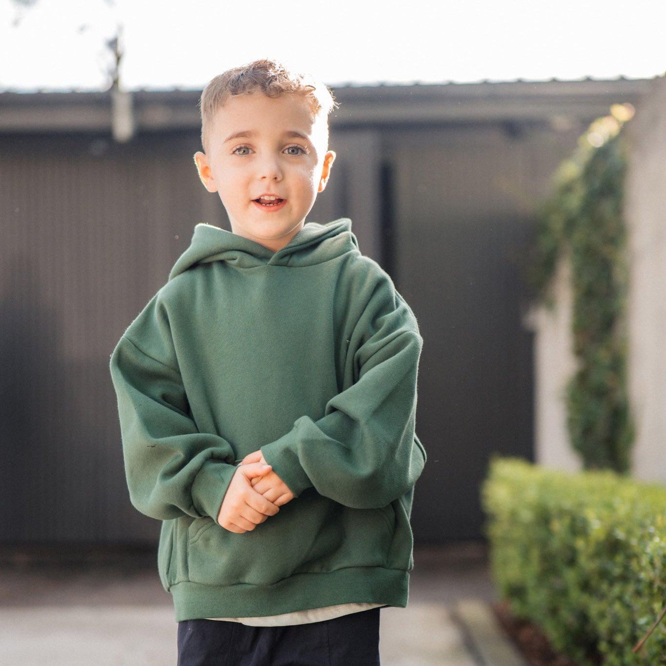 Child wearing a green hoodie and dark pants standing on a sidewalk.