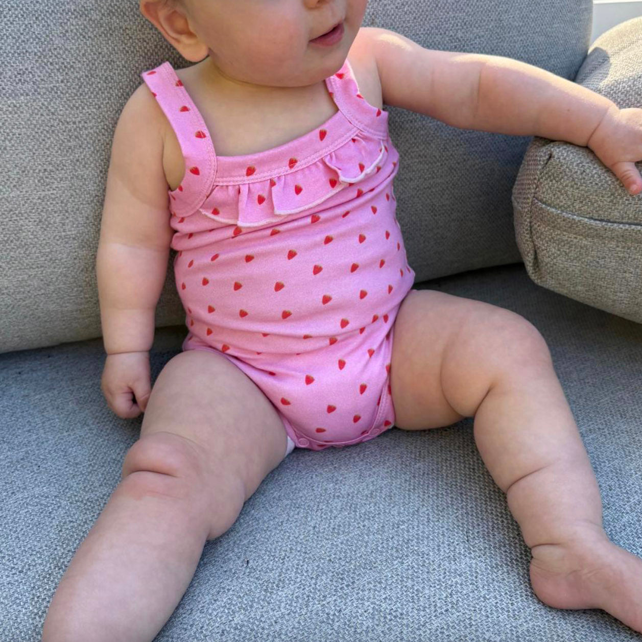 Baby wearing a pink playsuit with red strawberries sitting on a grey couch.
