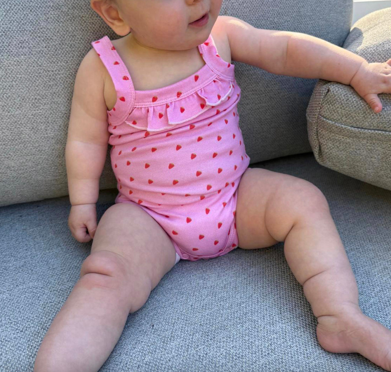 Baby wearing a pink playsuit with red strawberries sitting on a grey couch.