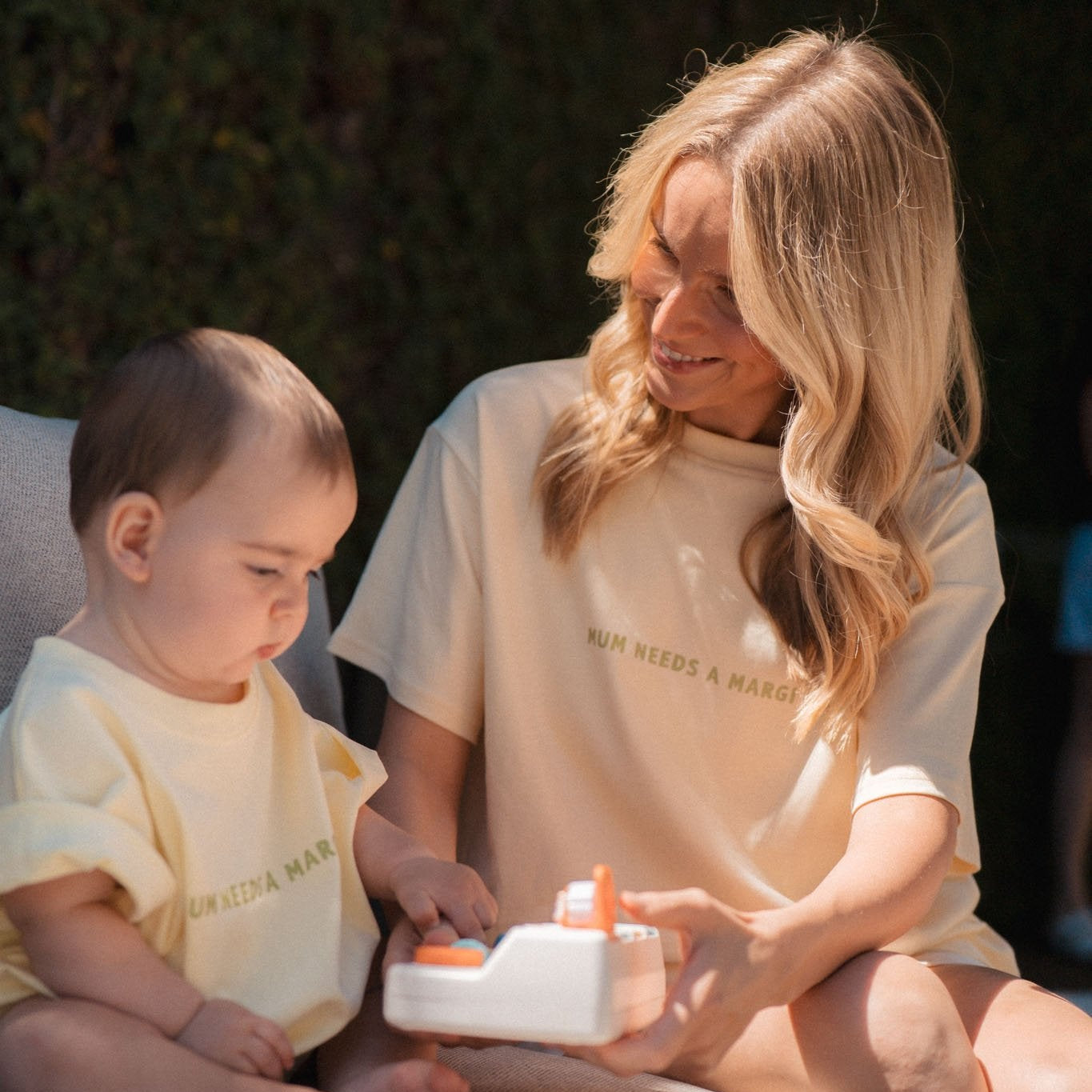 Woman and child sitting outdoors, both wearing matching light-colored t-shirts.